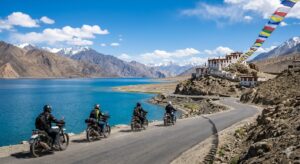 "लद्दाख की नीली पैंगोंग झील के किनारे सड़क पर बाइक चलाते चार राइडर्स, बैकग्राउंड में बर्फ से ढके पहाड़, एक बौद्ध मठ और हवा में लहराती रंग-बिरंगी बौद्ध प्रार्थना पताकाएं (Prayer Flags)."