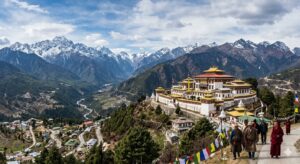 अरुणाचल प्रदेश में बर्फ से ढके हिमालय के पहाड़ों और हरी-भरी घाटी के बीच स्थित भव्य तवांग मठ का एक विस्तृत पैनोरमा दृश्य, जहां रास्ते पर पारंपरिक कपड़ों में बौद्ध भिक्षु और लोग घूम रहे हैं और रंग-बिरंगे प्रार्थना ध्वज लहरा रहे हैं।